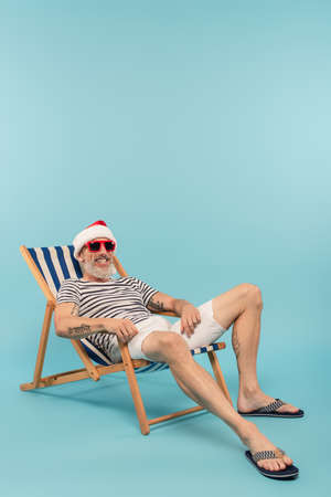 happy mature man in santa hat and sunglasses sitting on deck chair on blueの写真素材