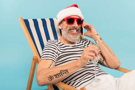 cheerful mature man in santa hat holding glass of champagne while talking on smartphone and sitting on deck chair isolated on blueの写真素材