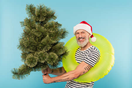 happy middle aged man in santa hat and swim ring holding christmas tree isolated on blue