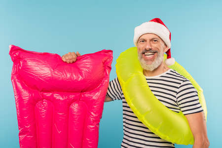 happy middle aged man in santa hat and swim ring holding pink inflatable mattress isolated on blueの写真素材