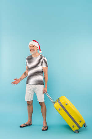 positive middle aged man in santa hat standing with luggage on blue