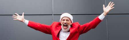 excited mature man in santa hat standing with outstretched hands near grey wall, bannerの写真素材