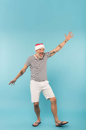 full length of excited man in santa hat standing with outstretched hands on blueの写真素材