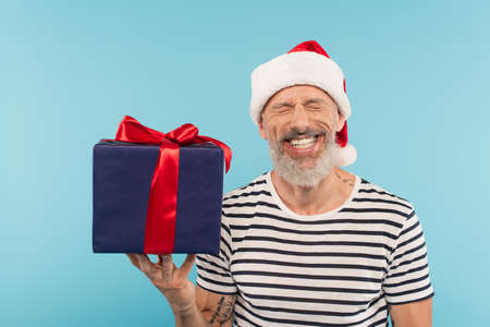 happy man with closed eyes in santa hat holding gift box isolated on blueの写真素材