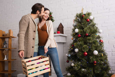 cheerful pregnant woman with husband holding wooden box with baubles near christmas treeの写真素材