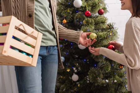 cropped view of man with wooden box giving christmas ball to wife decorating pine treeの写真素材