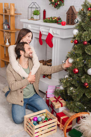 cheerful woman hugging husband decorating christmas tree in living roomの写真素材