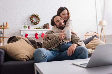 joyful woman hugging husband sitting on couch with cup of tea near blurred laptopの写真素材