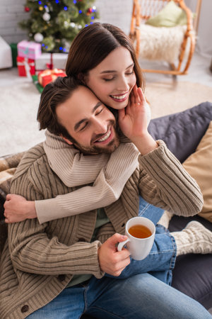 pleased man with cup of tea touching face of wife embracing him in living room with christmas decorationの写真素材