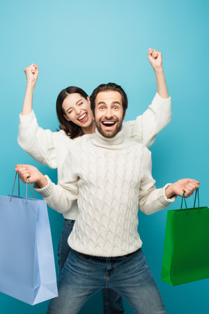 astonished man holding shopping bags near woman showing yeah gesture isolated on blueの写真素材