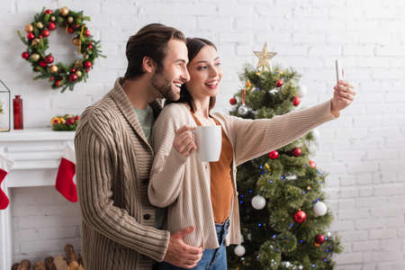 cheerful woman holding cup of tea while taking selfie with husband near blurred christmas treeの写真素材