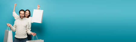 excited woman showing win gesture near man with shopping bags on blue, bannerの写真素材