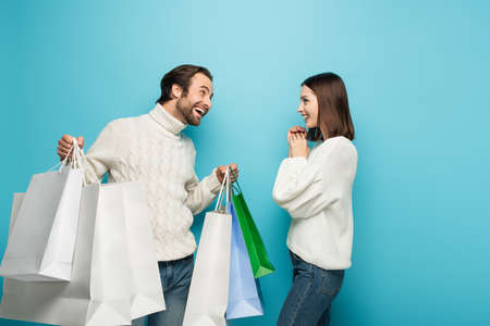 cheerful man holding shopping bags near amazed woman on blue