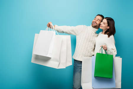 smiling couple in white sweaters looking away while holding purchases on blue