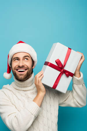happy and curious man in santa hat shaking gift box isolated on blue