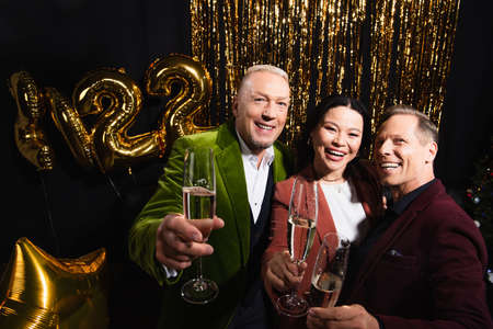 Smiling multiethnic friends holding champagne near balloons and tinsel on black background