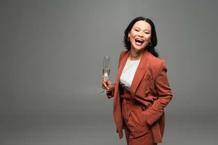 Excited asian woman in jacket holding champagne and looking at camera isolated on greyの写真素材