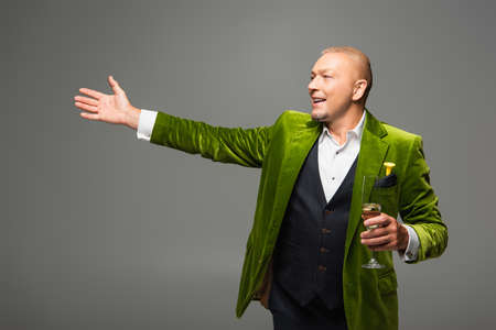 Positive man with glass of champagne pointing with hand and looking away isolated on grey