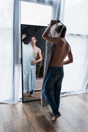 full length of young transgender man with makeup holding hanger with slip dress near mirrorの写真素材