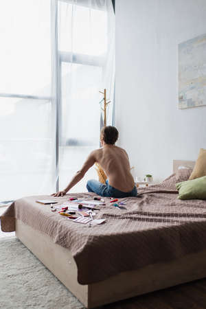 back view of shirtless young transgender man sitting on bed near makeup products
