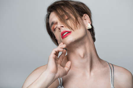 young transgender man with makeup and earring posing with closed eyes isolated on greyの写真素材