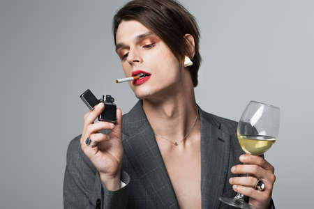 young transgender man in blazer holding glass of wine and lighter while smoking isolated on grey