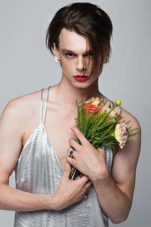 young transgender man in slip dress holding flowers isolated on grayの写真素材