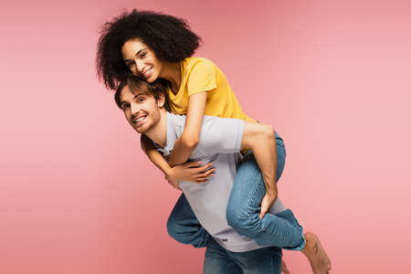 young and happy multiethnic couple smiling at camera while having fun isolated on pink