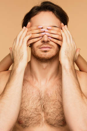 shirtless man touching hands of young woman obscuring his eyes isolated on beige