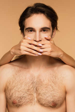 woman obscuring mouth of positive shirtless man looking at camera isolated on beigeの写真素材