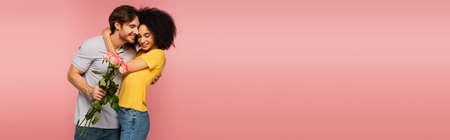 young man embracing happy hispanic woman while holding flowers isolated on pink, bannerの写真素材