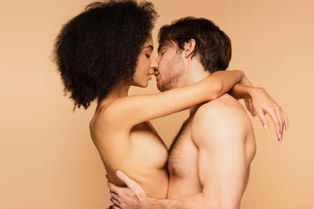 side view of sexy multiethnic lovers embracing and kissing with closed eyes isolated on beigeの写真素材