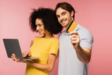 smiling hispanic woman using laptop near cheerful man holding credit card isolated on pinkの写真素材