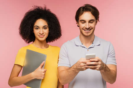 cheerful man chatting on smartphone near latin woman holding laptop isolated on pink