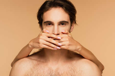young shirtless man looking at camera while woman covering his mouth with hands isolated on beigeの写真素材