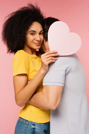happy latin woman with paper heart looking at camera while boyfriend embracing her isolated on pinkの写真素材