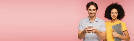 young hispanic woman with laptop smiling at camera near man chatting on smartphone isolated on pink, bannerの写真素材