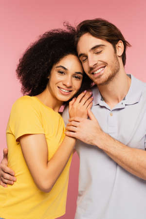 happy man with closed eyes hugging pretty hispanic woman looking at camera isolated on pinkの写真素材
