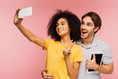 amazed man with digital tablet hugging hispanic girlfriend showing victory sign while taking selfie...