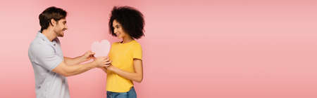 young man gifting paper heart to joyful latin woman isolated on pink, bannerの写真素材