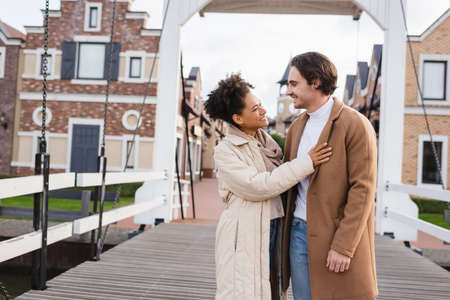 smiling multiethnic couple in coats hugging near shopping center