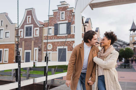 joyful multiethnic couple in coats holding hands and smiling near shopping centerの写真素材