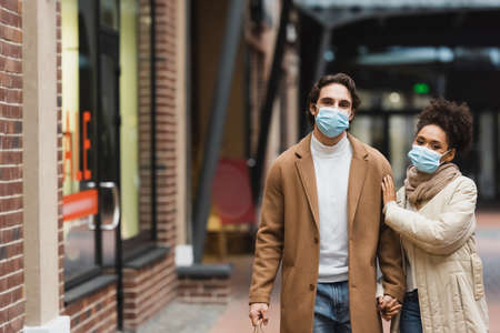 young multiethnic couple in medical masks holding hands while walking in shopping mallの写真素材