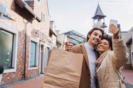 happy african american woman taking selfie with boyfriend holding shopping bags
