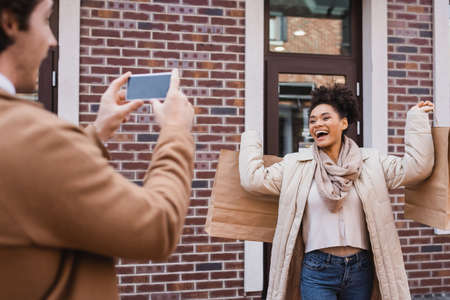 blurred man taking photo of excited african american woman with shopping bagsの写真素材