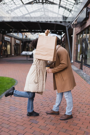 brunette african american woman holding shopping bags while covering faces of her and boyfriend in mallの写真素材