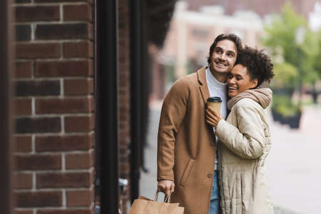 joyful african american woman holding paper cup near happy boyfriend with shopping bagsの写真素材
