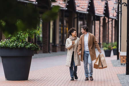 happy multiethnic couple in coats holding hands while walking with purchases near mallの写真素材