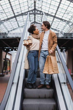 happy african american woman holding paper cups and looking at man with shopping bags on escalator...