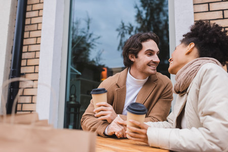 joyful interracial couple in coats holding hands and paper cups near blurred shopping bagsの写真素材